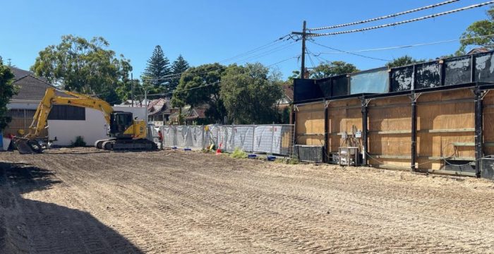 A construction site in Rose Bay with freshly leveled ground, an excavator parked on the left, and a fence separating the site from neighboring houses and structures. Trees and clear blue sky in the background hint at recent demolition in this picturesque Sydney suburb.