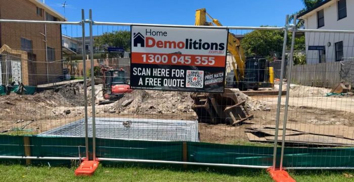A demolition site featuring a fence with a "Home Demolitions" sign stands ready, while construction machinery looms in the background. Measures for sediment control ensure the area remains secure and environmentally responsible amidst the ongoing work.