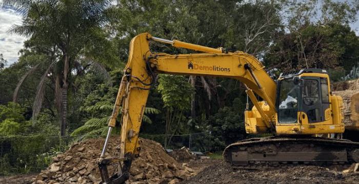 A yellow excavator is parked on a dirt construction site near piles of rocks and soil, surrounded by trees under a partly cloudy sky.
