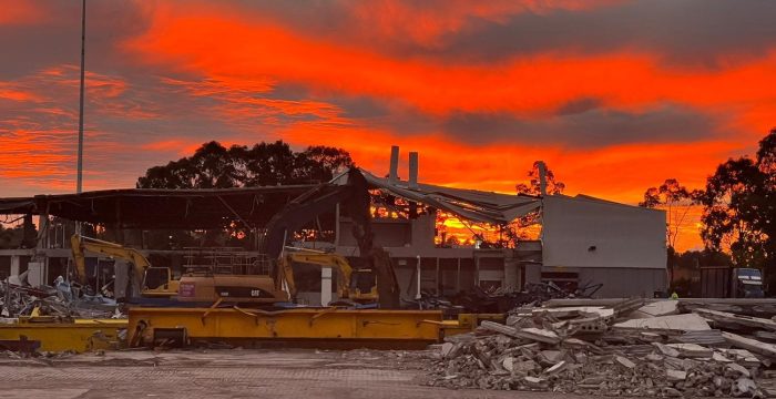 A construction site at sunset, featuring a partially demolished building, construction equipment, and scattered debris under a vibrant orange sky reminiscent of sunrise.